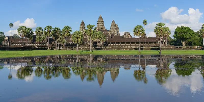 Las torres de Angkor Wat se alzan entre palmeras y se reflejan en el agua quieta de un estanque, bajo un cielo azul.