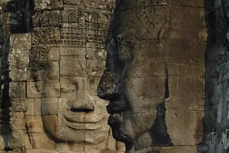 Dos enormes rostros de piedra tallados en las torres del Bayon, uno sonriente de frente y otro de perfil en sombra.