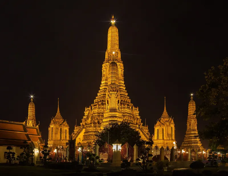 La torre central de Wat Arun y sus torres menores, iluminadas de dorado, contra el cielo negro de la noche.
