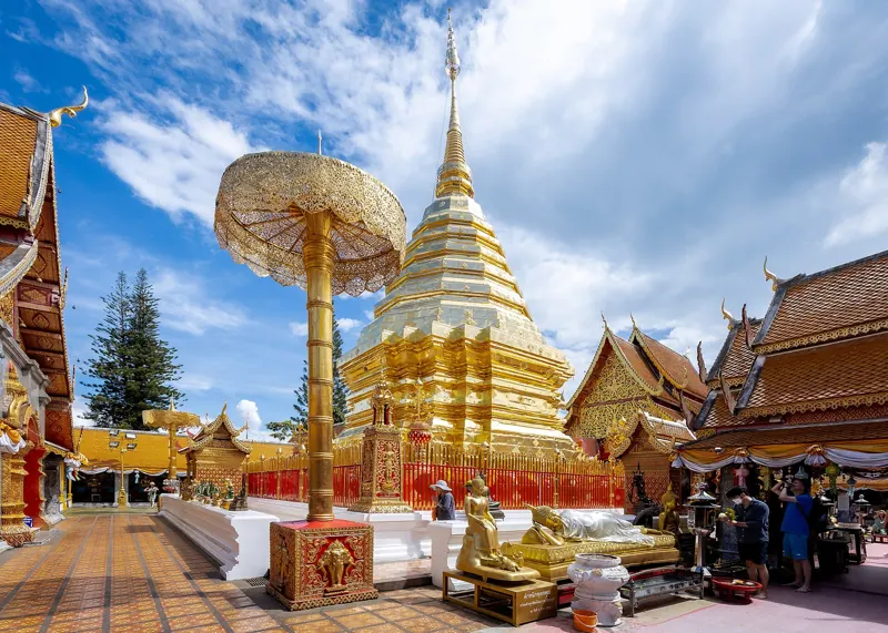 Chedi dorado de Wat Phra That Doi Suthep junto a un parasol ceremonial dorado, entre templos de techos rojizos y cielo azul.
