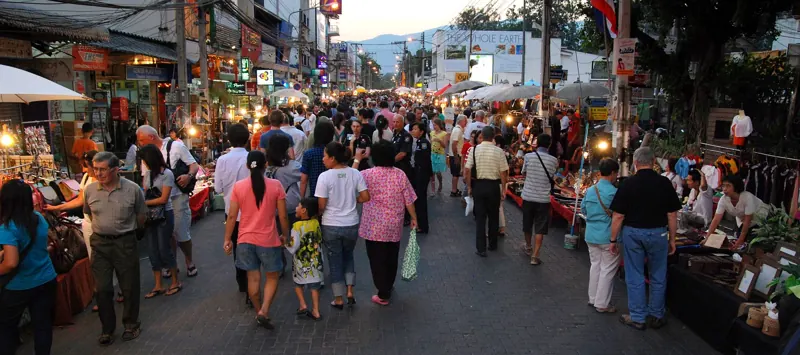 Multitud al anochecer en el Mercado Peatonal del Domingo de Chiang Mai, con puestos iluminados a ambos lados de la calle.