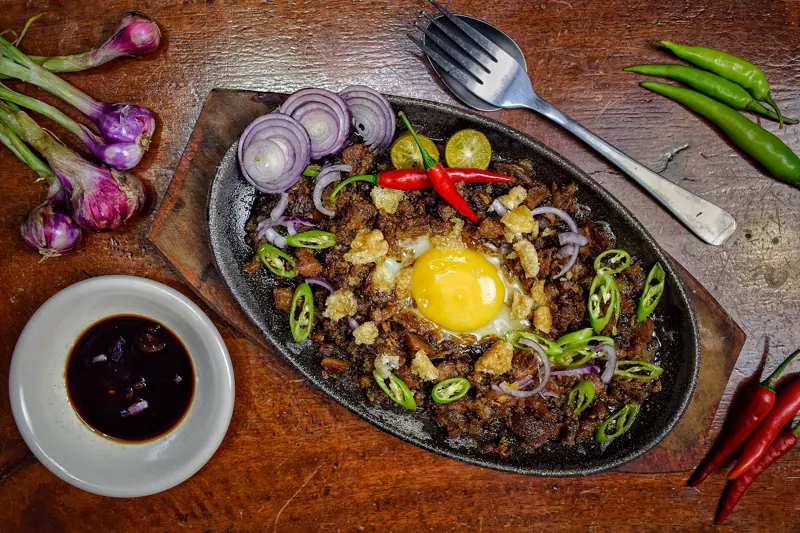 Sisig en un plato de hierro caliente, con una yema al centro, cebolla morada, ají verde y calamansí sobre una mesa de madera.