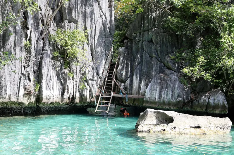 Escalera apoyada en una pared de roca gris sobre agua turquesa, con un bañista de chaleco naranja al pie.