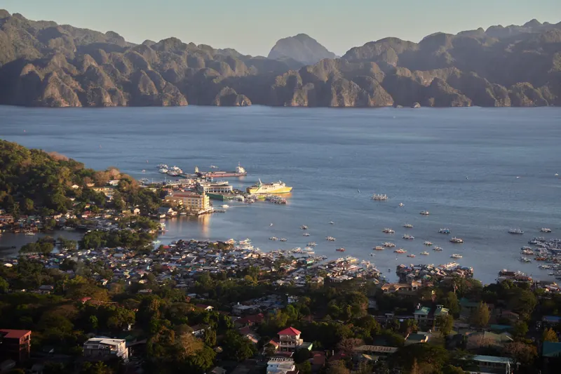 Pueblo y puerto de Coron vistos desde lo alto con luz de tarde, botes en la bahía y montañas de roca al fondo.