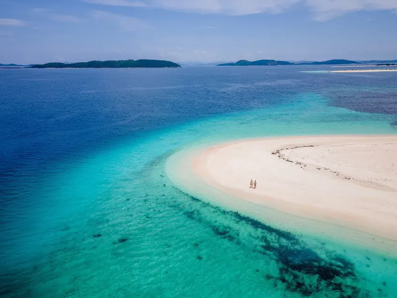 Vista aérea de un banco de arena blanca rodeado de agua turquesa y arrecifes, con dos personas diminutas e islas al fondo.