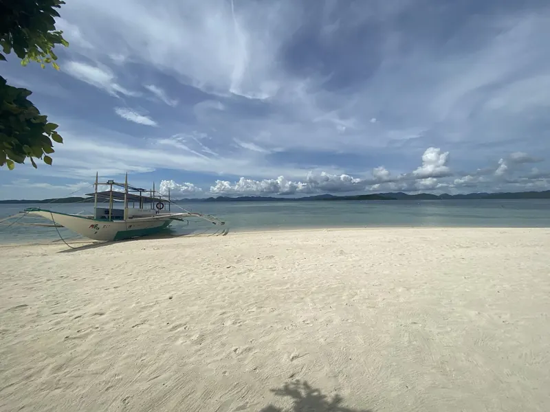 Bangka en la orilla de una playa de arena blanca y vacía, con agua turquesa e islas bajas bajo un cielo con nubes.