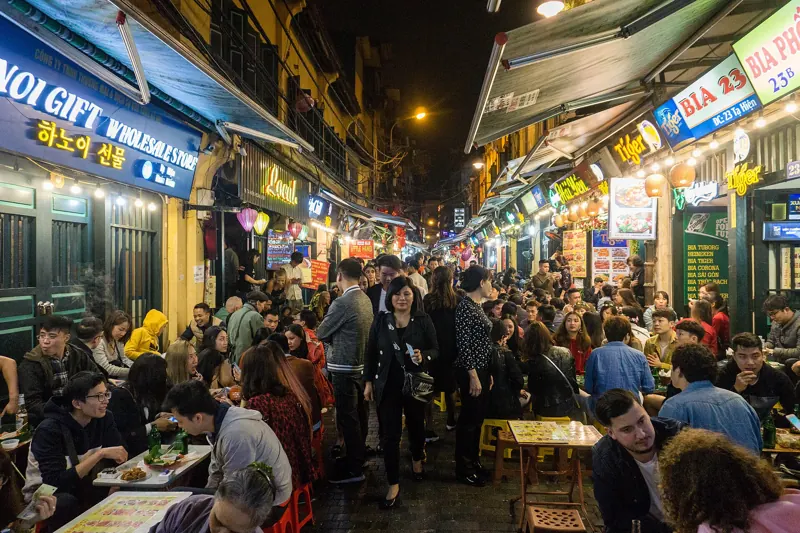 Calle del Barrio Antiguo de Hanói de noche, repleta de gente sentada en mesas bajas o caminando bajo letreros luminosos.