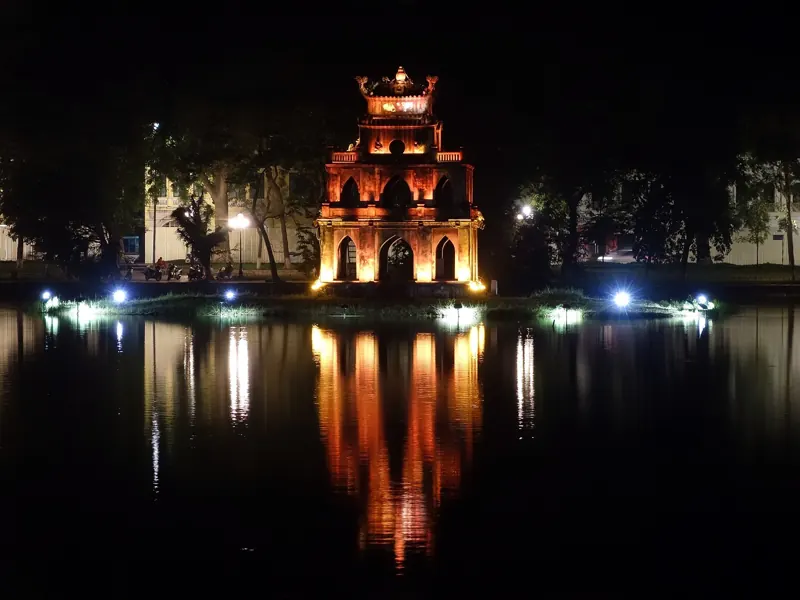 La Torre de la Tortuga iluminada de noche en medio del lago Hoan Kiem, con su reflejo naranja en el agua oscura.