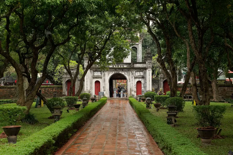 Camino de ladrillo entre setos y árboles que lleva a la puerta principal del Templo de la Literatura, con el suelo mojado.