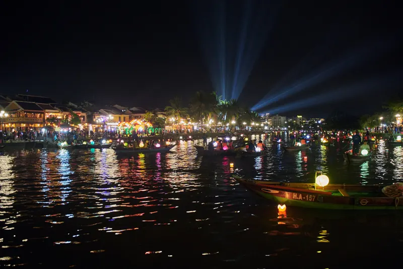 Botes con faroles y luces flotando en el río Thu Bon de noche, frente a la ribera iluminada de Hoi An, con haces de luz en el cielo.