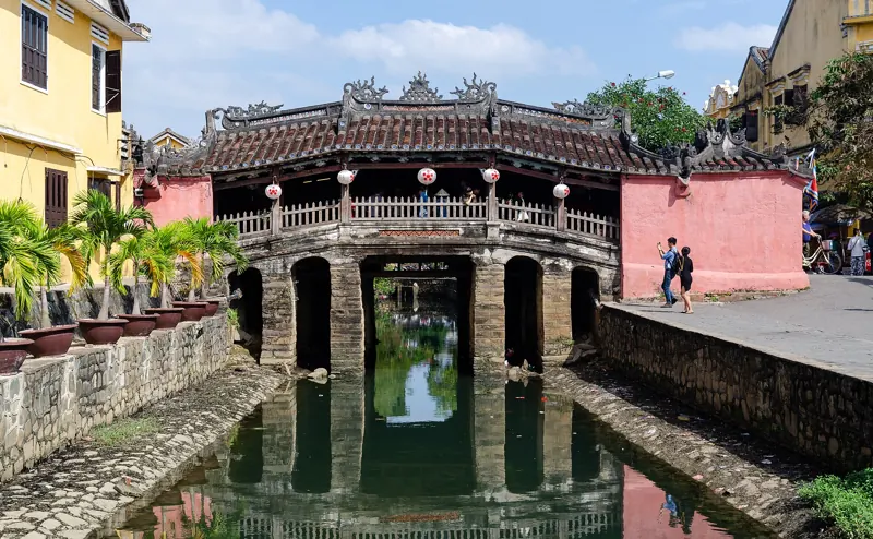 El Puente Japonés de Hoi An, con techo de tejas y pilares de piedra, reflejado en el agua del canal en un día soleado.