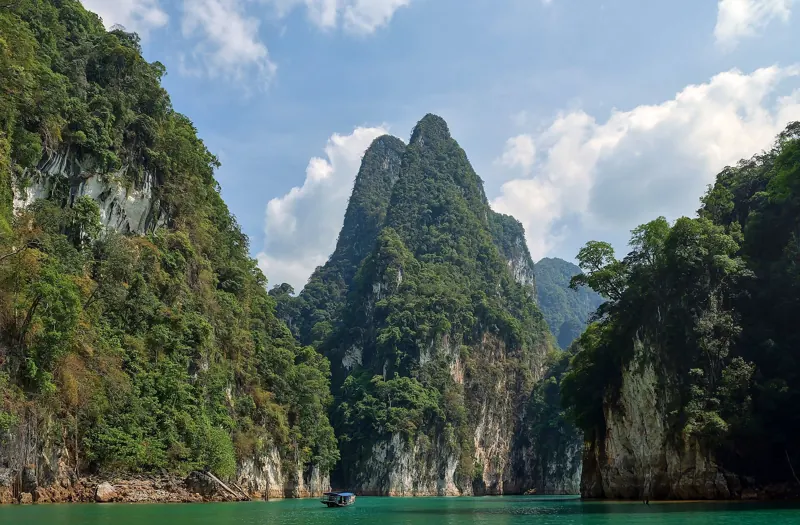 Paredes de karst cubiertas de selva se elevan sobre el agua verde del lago Cheow Lan; abajo navega un bote pequeño.