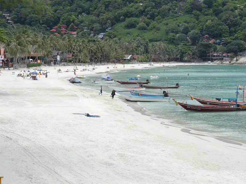 Playa de arena blanca de Haad Rin Nok de día, con botes longtail en la orilla y palmeras y cerros verdes detrás.