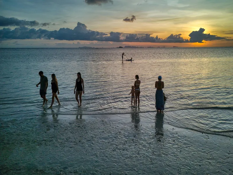 Siluetas de personas que caminan por el agua poco profunda de Koh Phangan al atardecer, con un remero de pie al fondo.