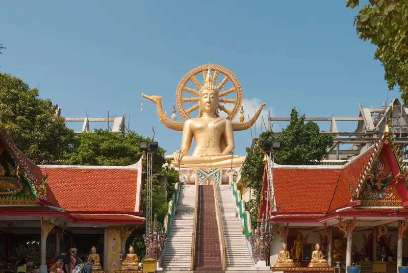 Gran Buda dorado sentado en lo alto de una escalinata, entre templos de techos rojos y bajo un cielo azul.