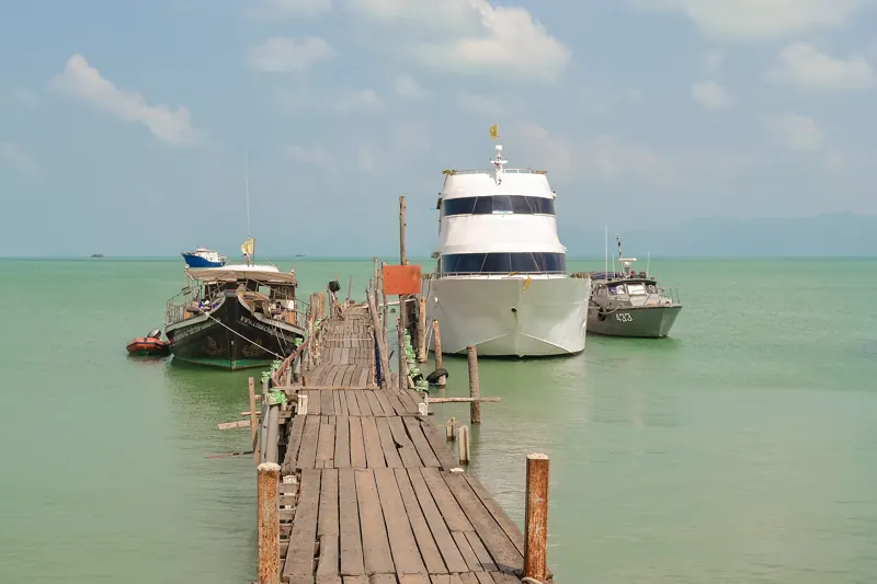 Muelle de madera que entra en el mar verde claro de Bophut, con un yate blanco y botes amarrados al final.