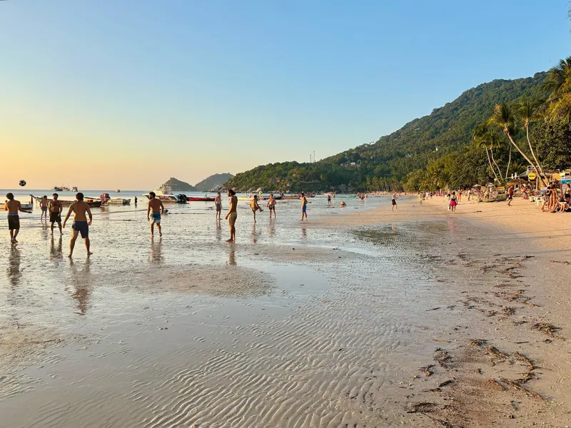 Gente camina y juega en la arena mojada de Sairee con marea baja al atardecer, con cerros verdes a la derecha.