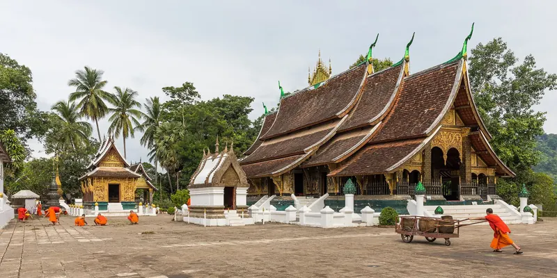 Monjes jóvenes de túnica naranja limpian el patio del Wat Xieng Thong, un templo de techos escalonados que bajan casi al suelo.