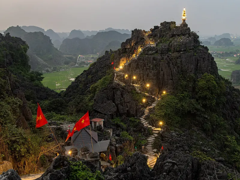 Escalera de piedra iluminada que sube entre rocas calizas hasta la pagoda de la cima de Hang Mua, al anochecer.