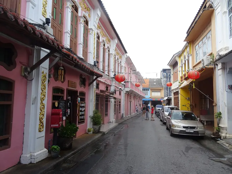 Calle Soi Romanee con casas-tienda sino-portuguesas rosadas y amarillas y faroles rojos colgados sobre la calle mojada.