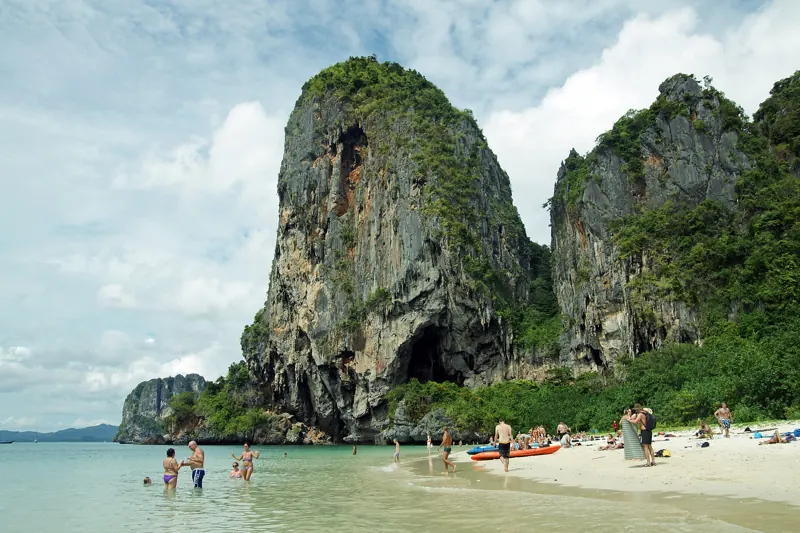 Un enorme peñón de roca caliza, con una cueva en la base, se alza sobre la playa de Phra Nang y su agua clara con bañistas.