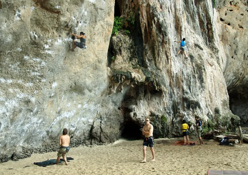Dos escaladores suben por una pared de roca caliza en la playa de Phra Nang; abajo, en la arena, otros los aseguran con la cuerda.