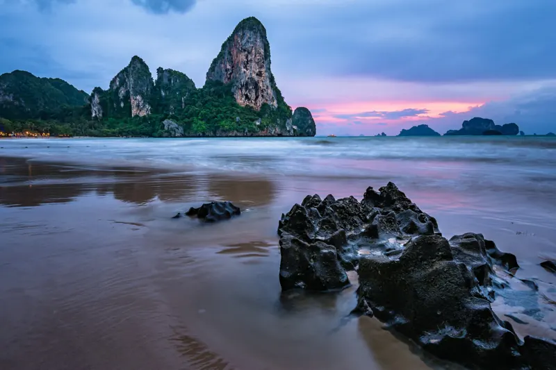 Railay West al anochecer: un peñón de karst sobre el mar, el horizonte rosado y rocas oscuras en la arena mojada.