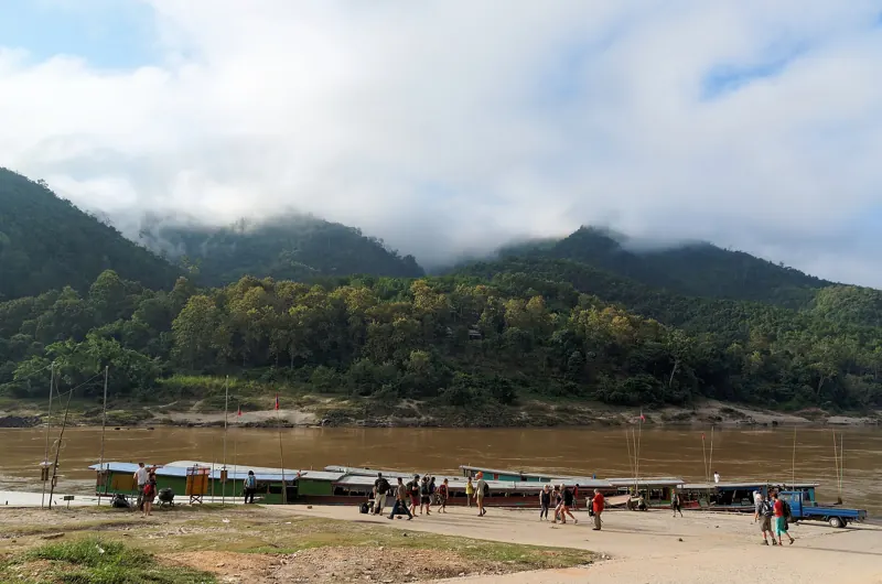 Botes largos y techados amarrados a la orilla del Mekong en Pakbeng, con viajeros caminando y cerros de selva entre nubes.