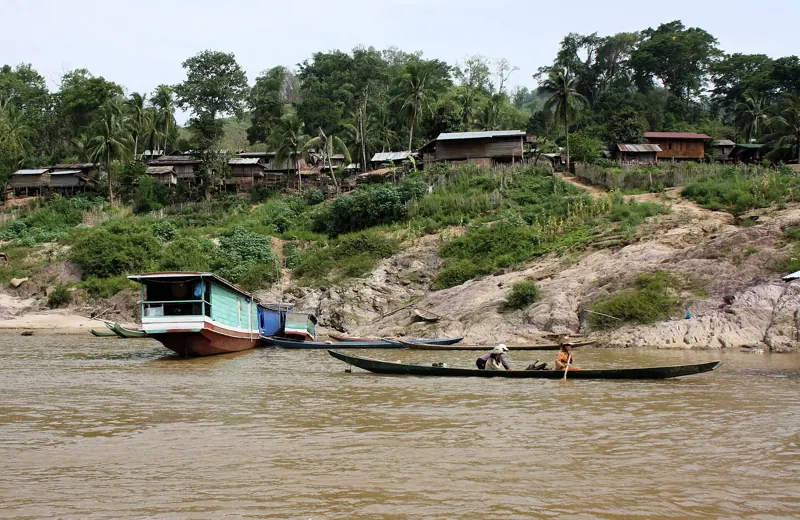 Un bote de pasajeros junto a la orilla y una canoa con dos personas en el Mekong, bajo una aldea de casas de madera.
