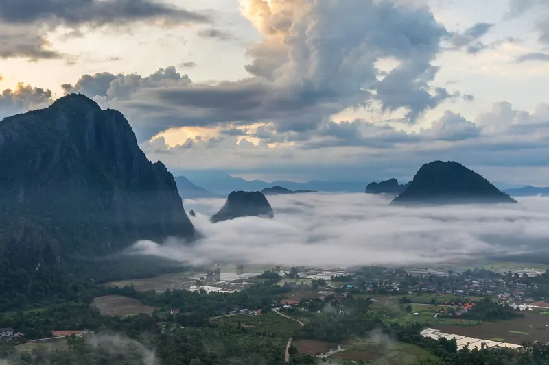 Picos de karst sobresalen de un mar de nubes al amanecer, con los campos y casas de Vang Vieng abajo.