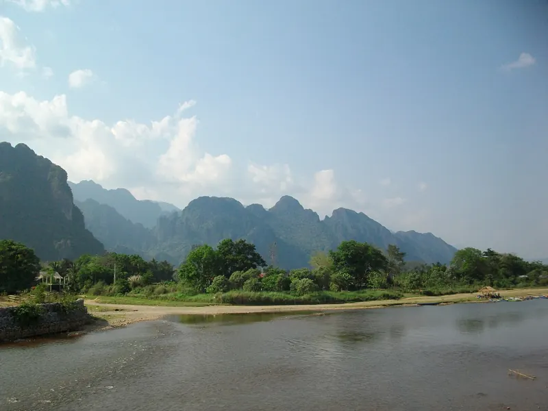 El río Nam Song corre ancho y tranquilo junto a una orilla de arena, bajo una hilera de montañas de karst.