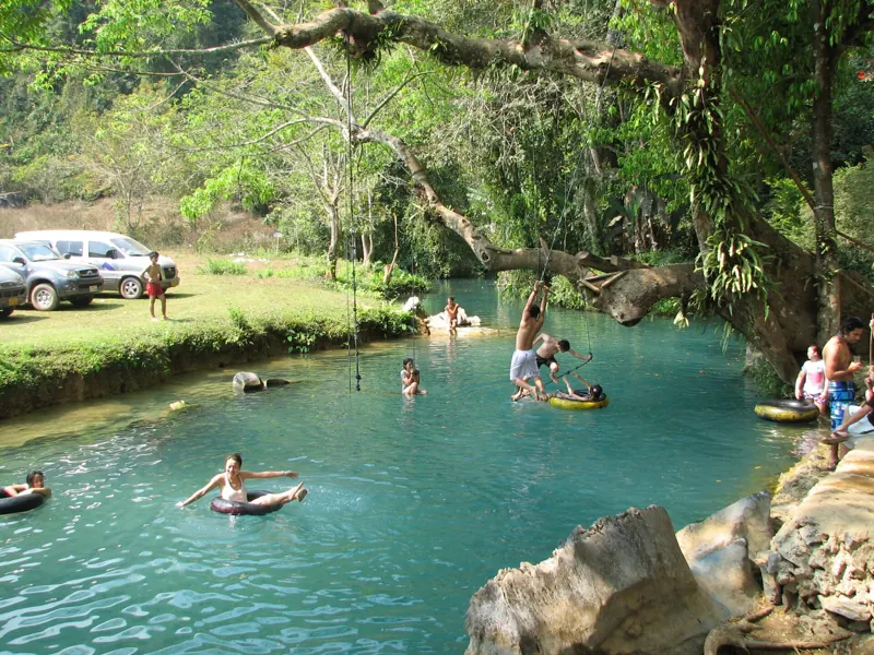 Bañistas flotan en neumáticos en el agua turquesa de la Laguna Azul, bajo un gran árbol del que cuelga una cuerda.